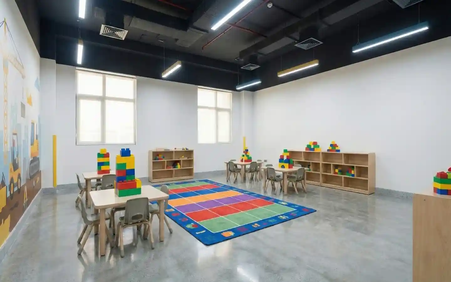 Modern preschool classroom featuring wooden activity tables, colorful building blocks, a checkered play rug, and industrial-style exposed ceilings