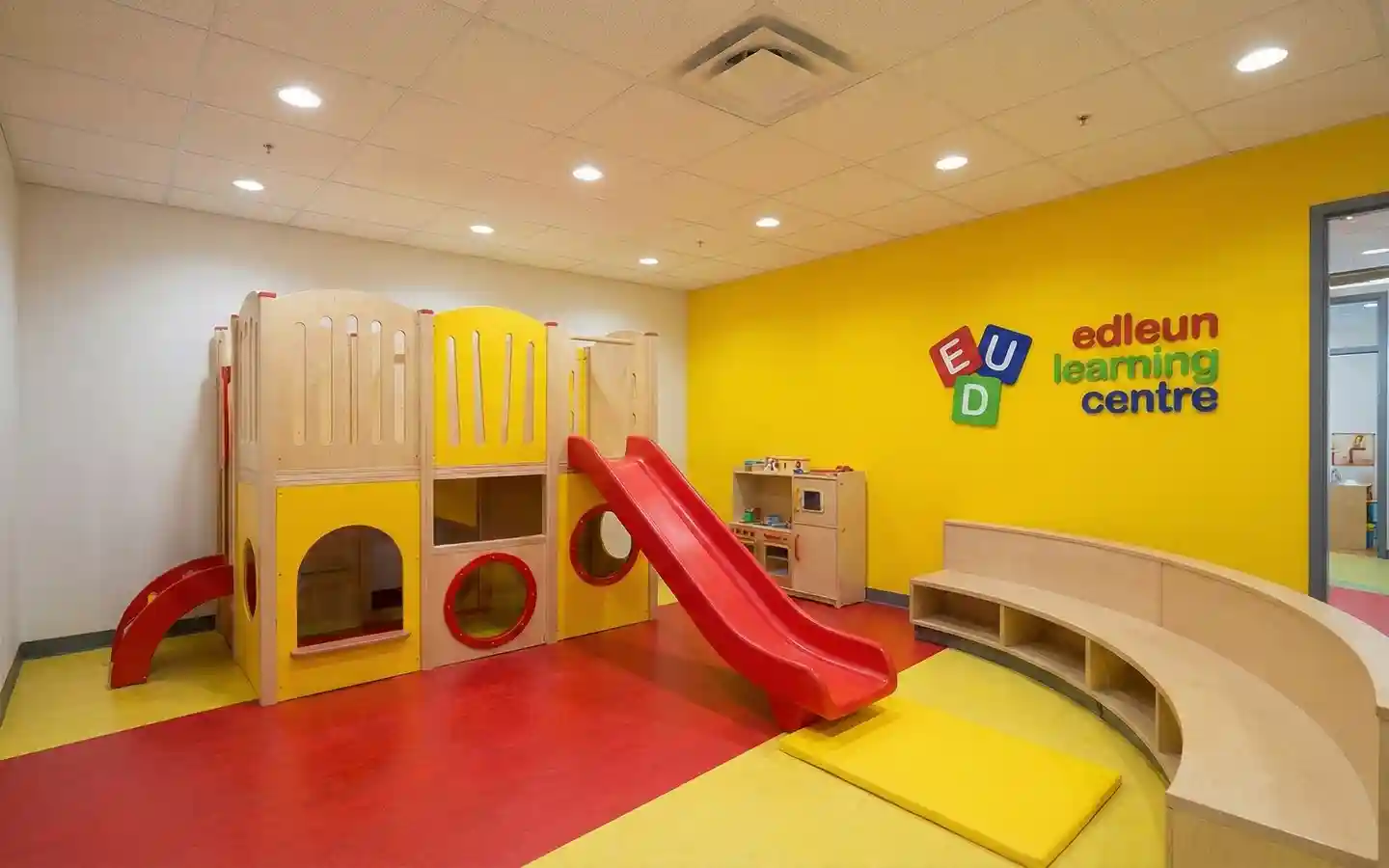 Vibrant preschool indoor play area featuring a wooden play structure with a red slide, yellow accent wall, and curved wooden seating at Edleun Learning Centre