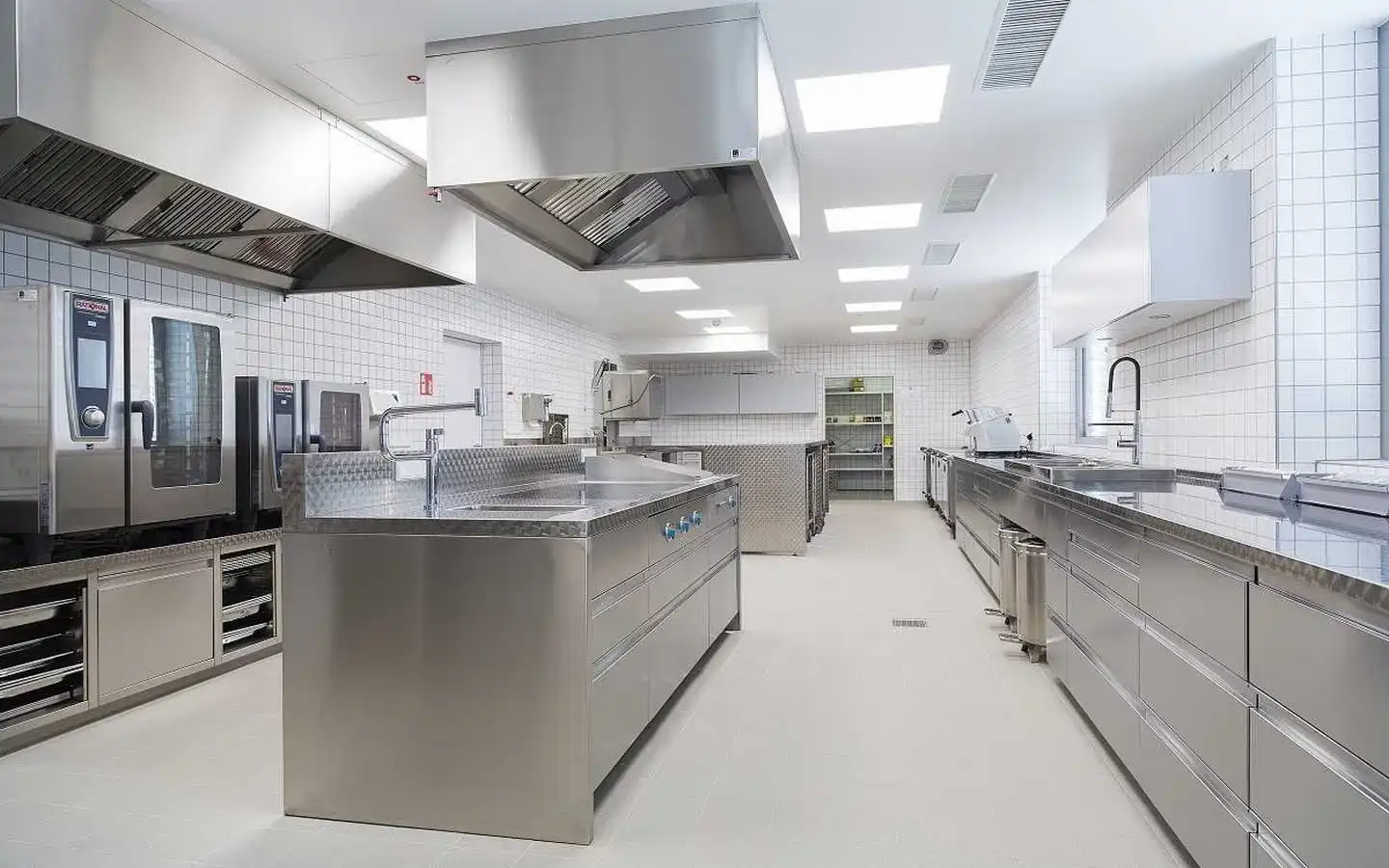 Professional commercial kitchen interior featuring industrial stainless steel workstations, large exhaust hoods, professional ovens, and white tiled walls