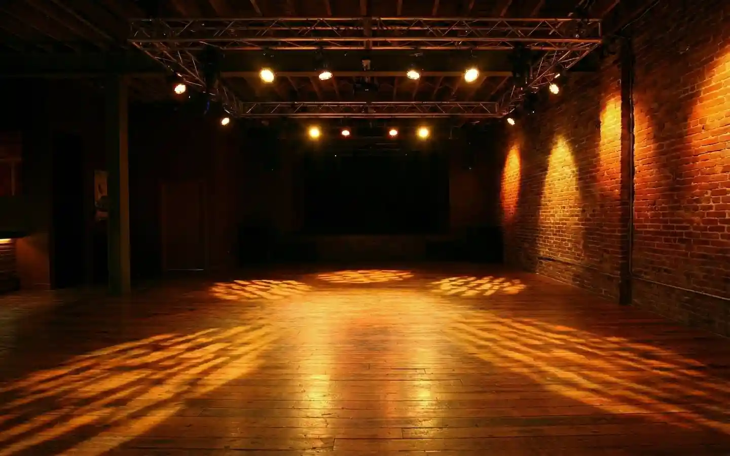 Industrial pub event venue with exposed red brick walls, hardwood floors, and professional stage lighting truss with warm spotlights