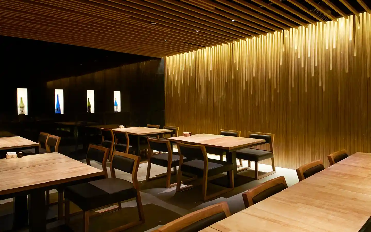 Upscale restaurant interior with dramatic wood slat wall lighting, timber ceiling baffles, and minimalist wooden dining furniture