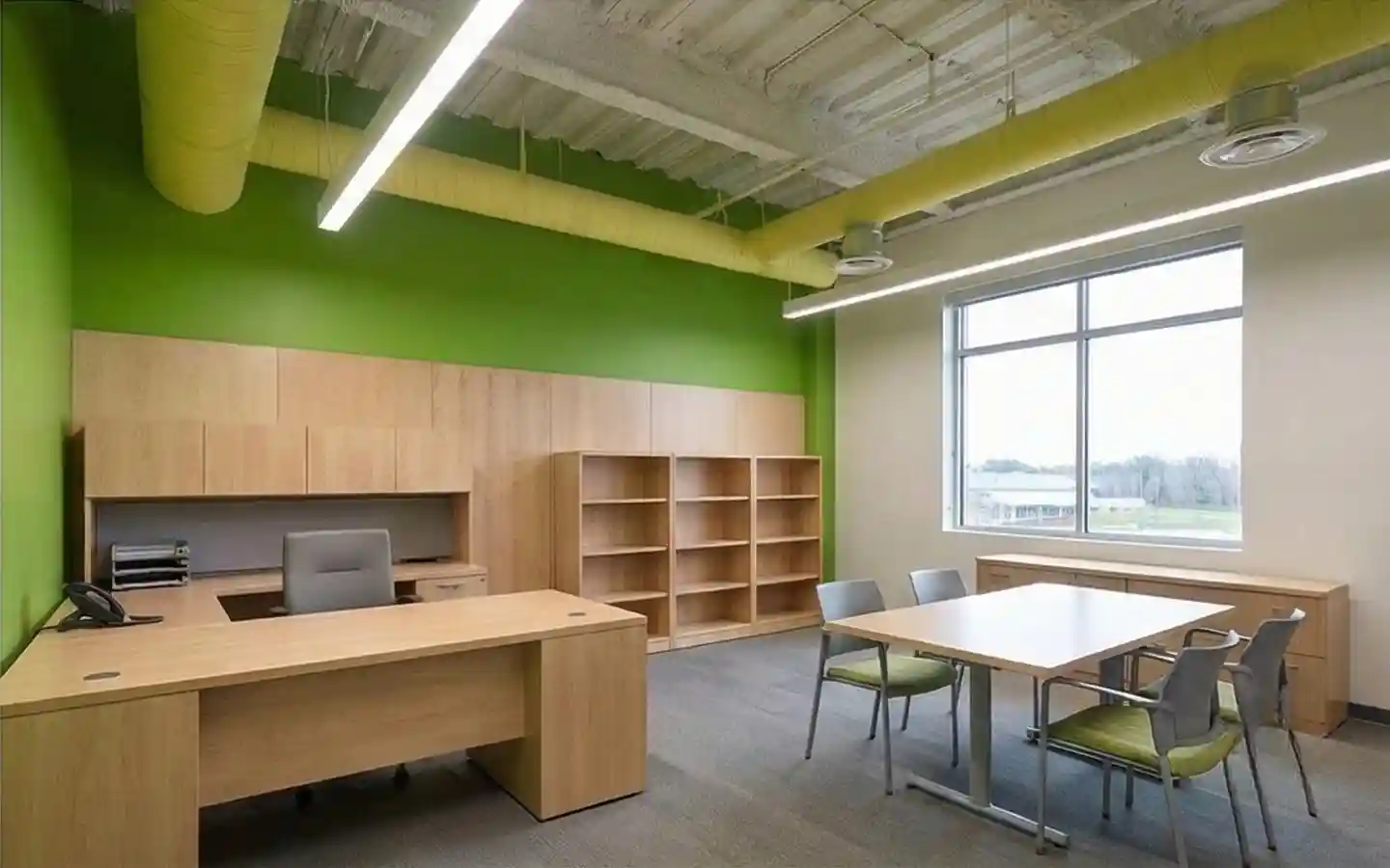Modern school administrative office featuring light wood executive desks, matching bookshelves, a green accent wall, and exposed industrial ceiling pipes