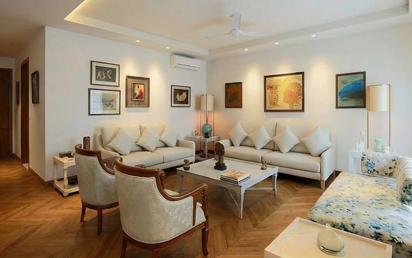 Spacious living room design featuring symmetrical cream sofas, a gallery wall with framed art, vintage wooden chairs, and warm recessed ceiling lighting over chevron parquet flooring