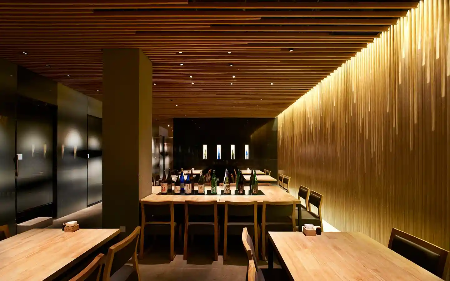 Restaurant interior featuring a warm wood slat ceiling, a vertical louvered feature wall with gold lighting, and a communal dining table with sake bottle display