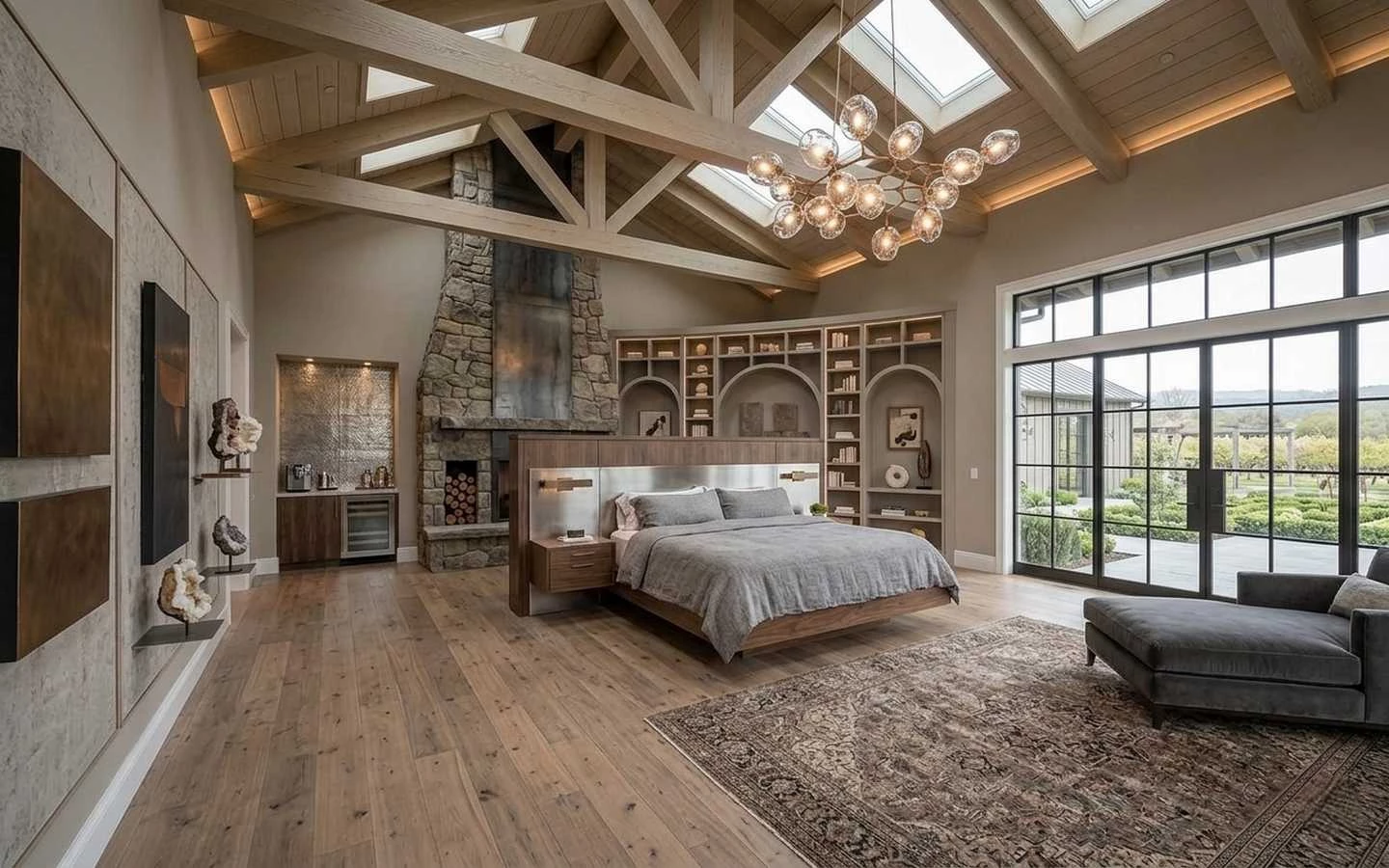 Luxury modern farmhouse master bedroom featuring a vaulted timber truss ceiling with skylights, a stone fireplace, integrated wet bar, and a floating bed design